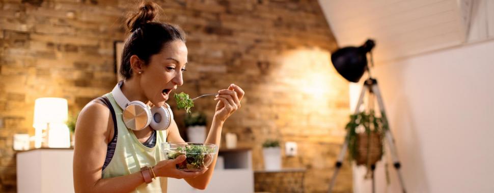 Mulher com headphone no pescoço comendo uma salada enquanto presta atenção na tela do computador. Crédito Pixabay